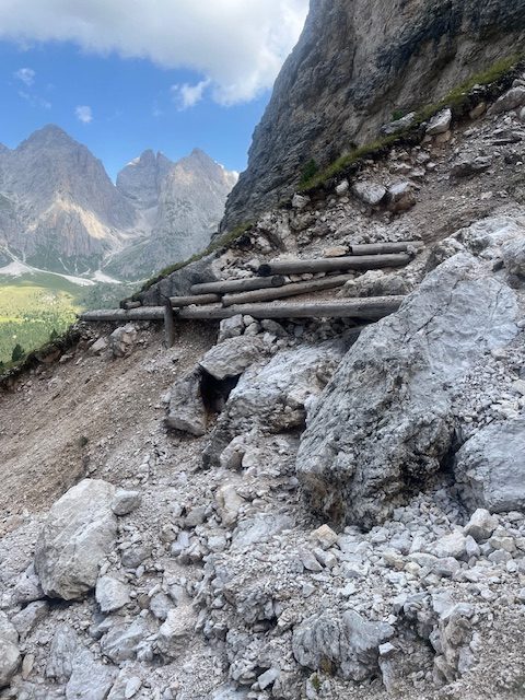 A weathered wooden walkway built across a steep scree slope, with jagged peaks in the distance