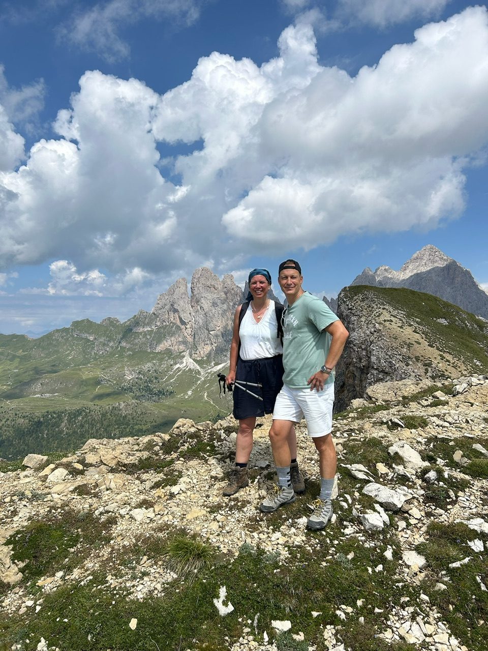 Camila and her husband standing together on a rocky summit in the Dolomites under a big summer sky